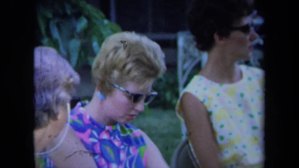1971 California 8mm frame of adult women in patterned sleeveless dresses sitting in a yard, with sunglasses and greenery visible.