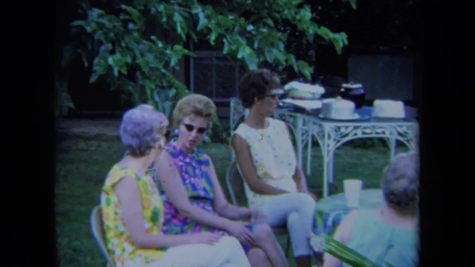 1971 California home-movie frame showing women seated outdoors in a yard, with a white metal table, cups or dishes, and leafy plants behind them.