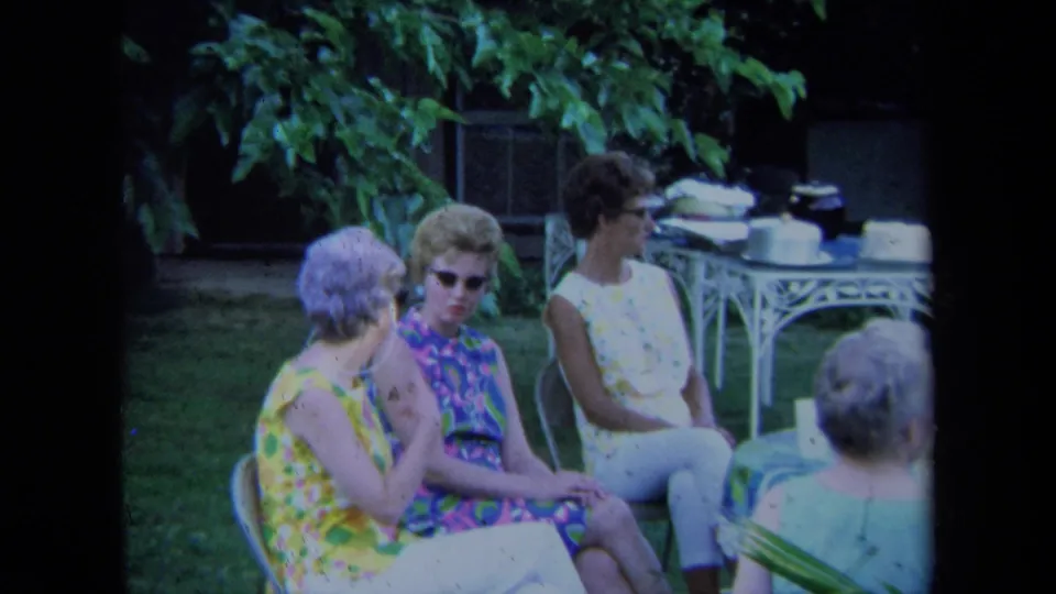 Adult women in a California yard appear mid-conversation, seated on lawn chairs with a white table and green foliage in the background.