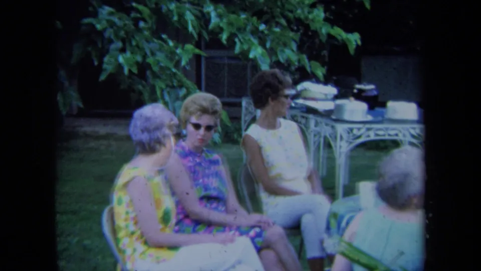 1971 California home-movie frame of women in colorful dresses and sunglasses seated at a garden gathering.