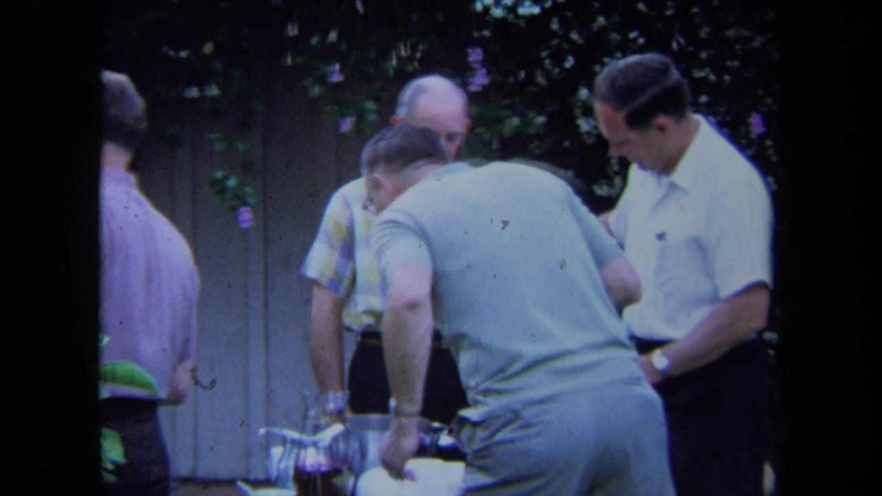 Adult men in short-sleeved shirts gather outdoors in California, 1971, near a table where a glass pitcher is being handled.