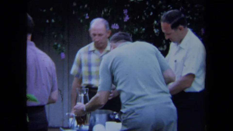 In a 1971 California garden setting, adult men stand close together while one leans over a table with a glass pitcher and cups.