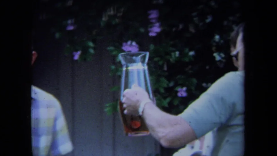 Close 1971 California home-movie view of an adult man's arm holding a glass pitcher with amber beer against garden foliage.
