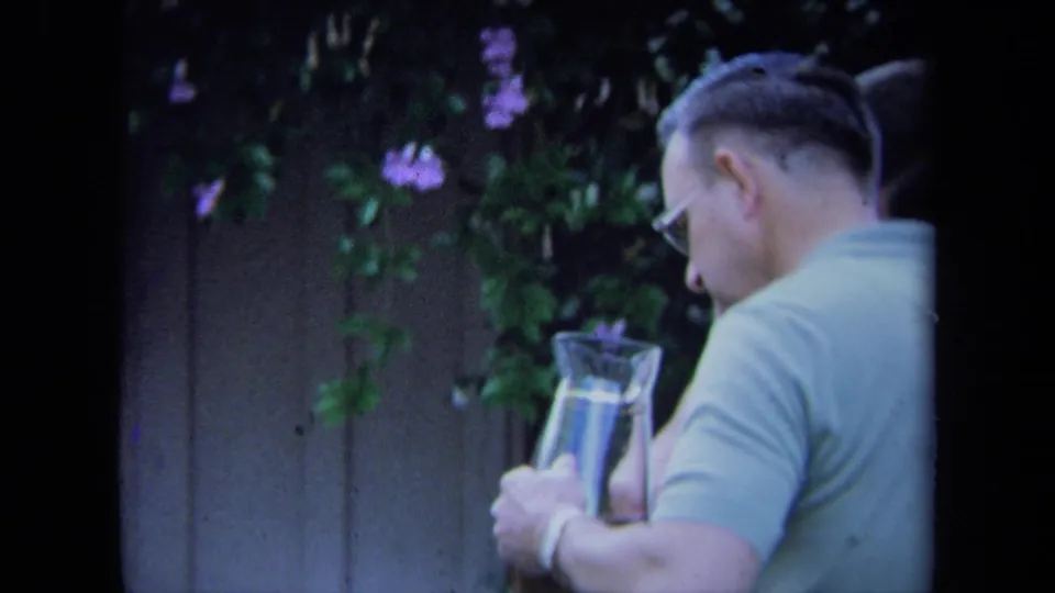 In a 1971 California backyard, a man in glasses holds a glass pitcher near flowering vines along a wooden fence.