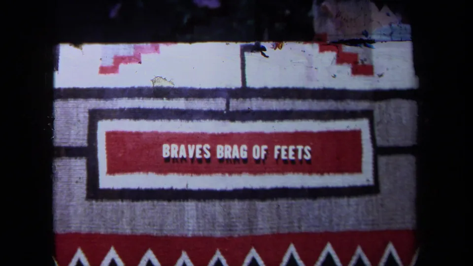 Close view of a woven textile with red geometric bands and a central panel reading BRAVES BRAG OF FEETS.