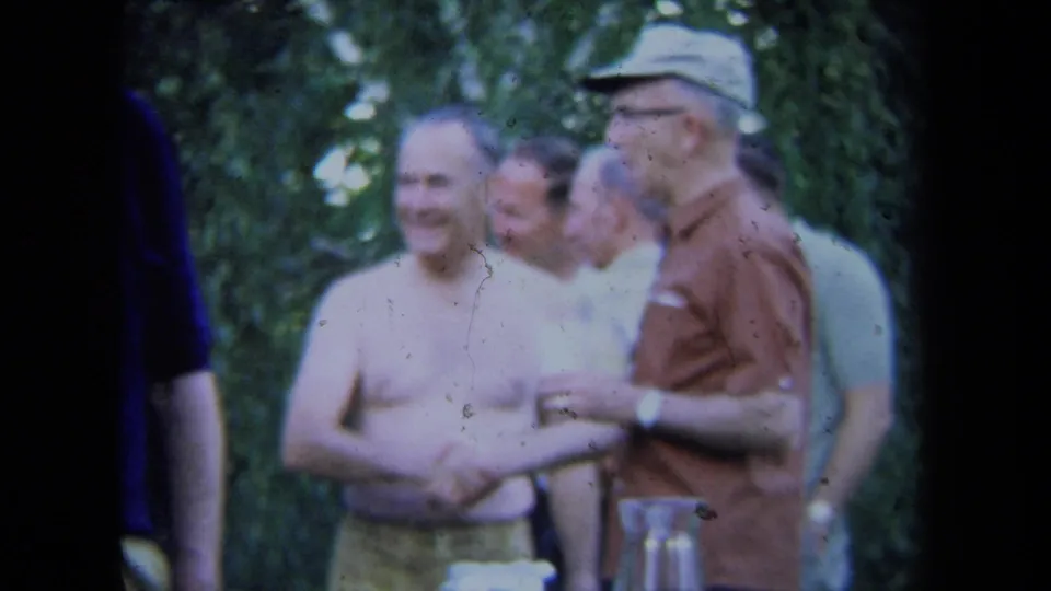 1971 California home-movie frame showing adult men laughing outdoors, including a shirtless man and a man in a brown shirt and cap.