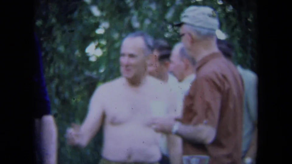 1971 California 8mm frame with a shirtless man smiling while speaking with a man in a brown shirt at an outdoor social event.
