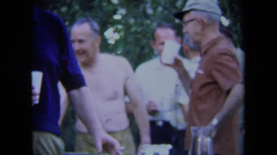 1971 California home-movie frame showing men gathered near paper cups and a glass pitcher, with one man lifting a cup.