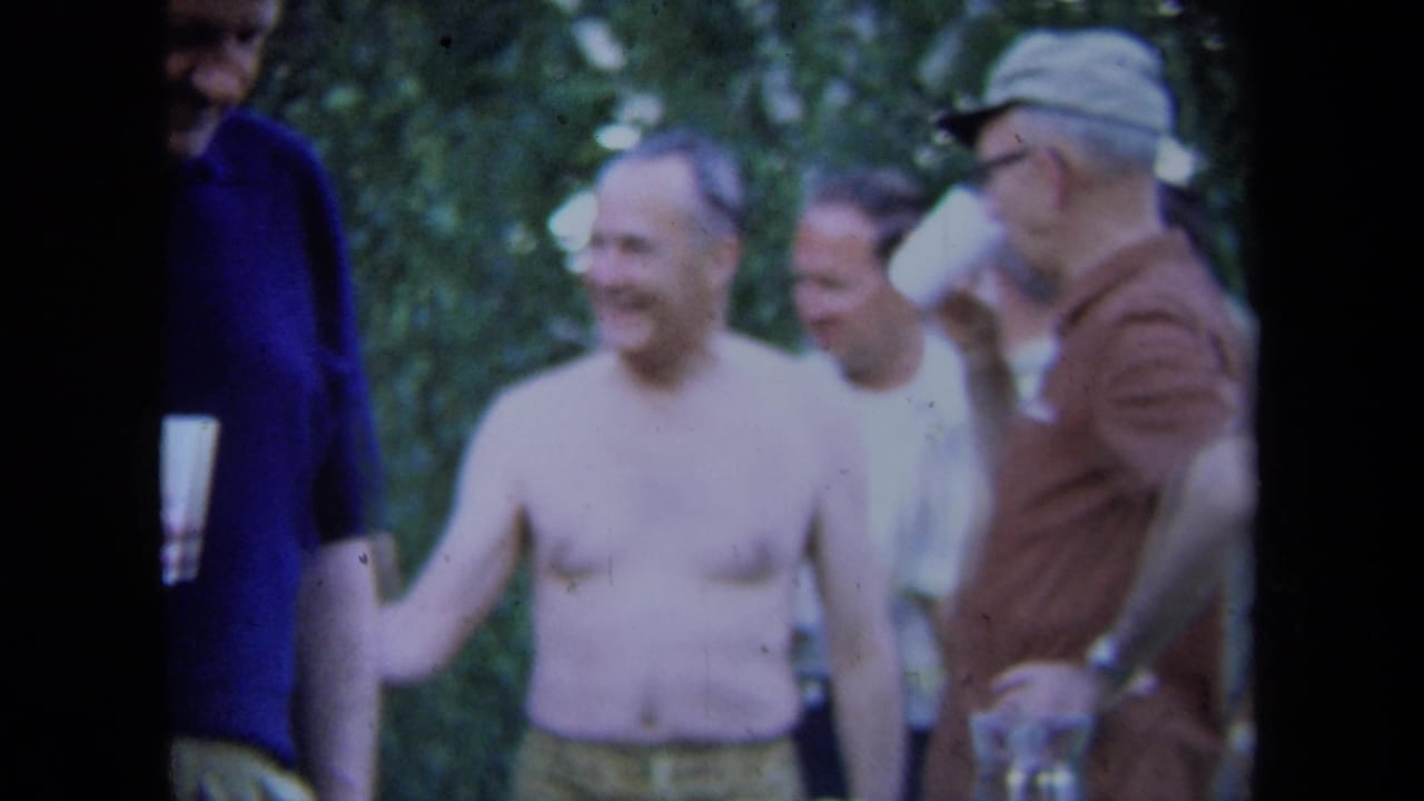 Adult men stand outdoors near trees, including one shirtless man and others holding white cups.