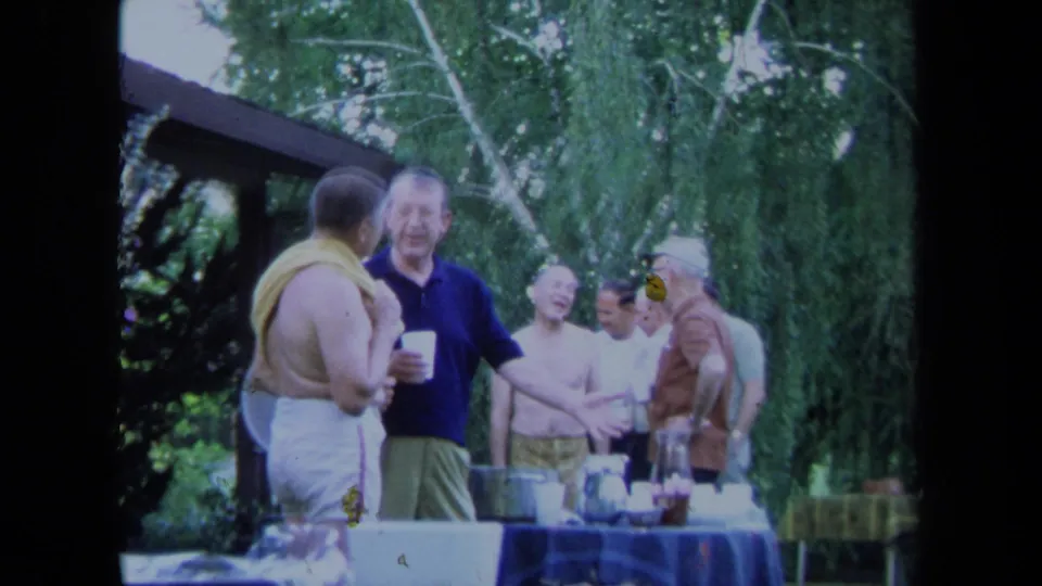1971 California home-movie frame of men gathered beside a blue-covered table with cups and serving items outdoors.