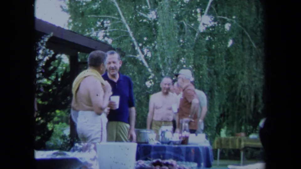 1971 California home-movie frame showing men socializing around a backyard table under dense green trees.