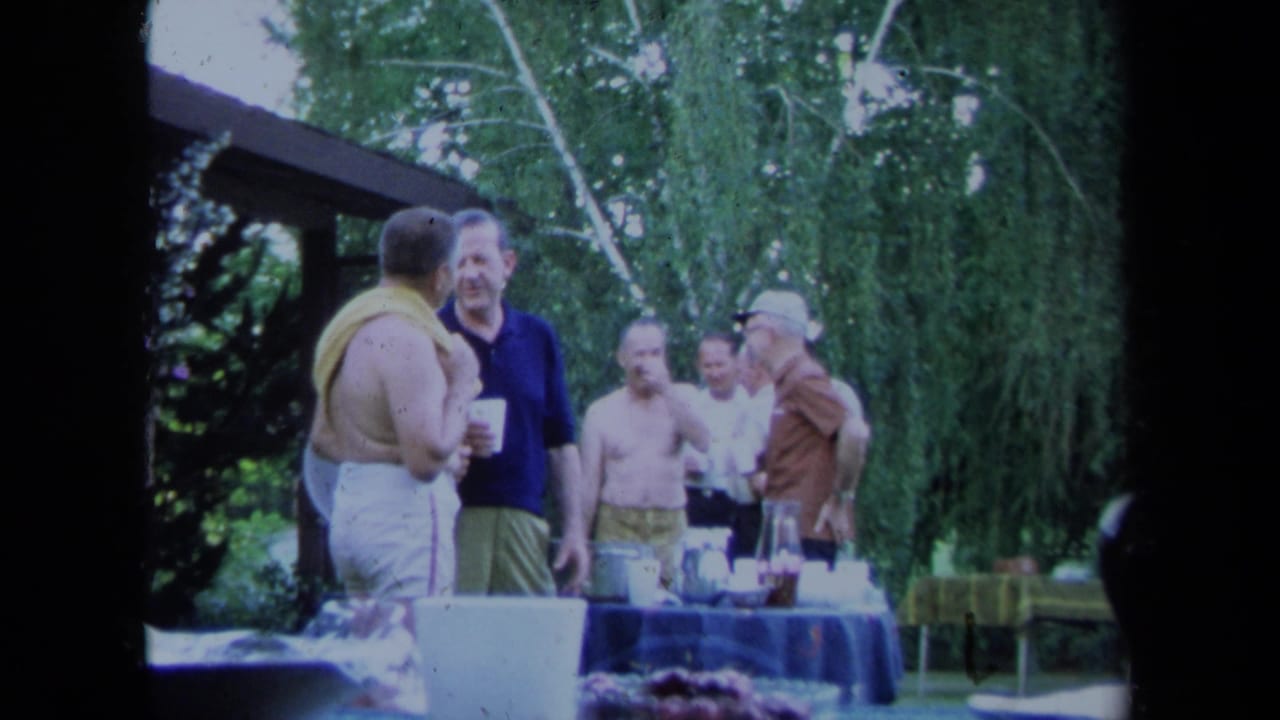 Adult men in 1971 California appear to talk beside an outdoor food and drink table in a garden setting.
