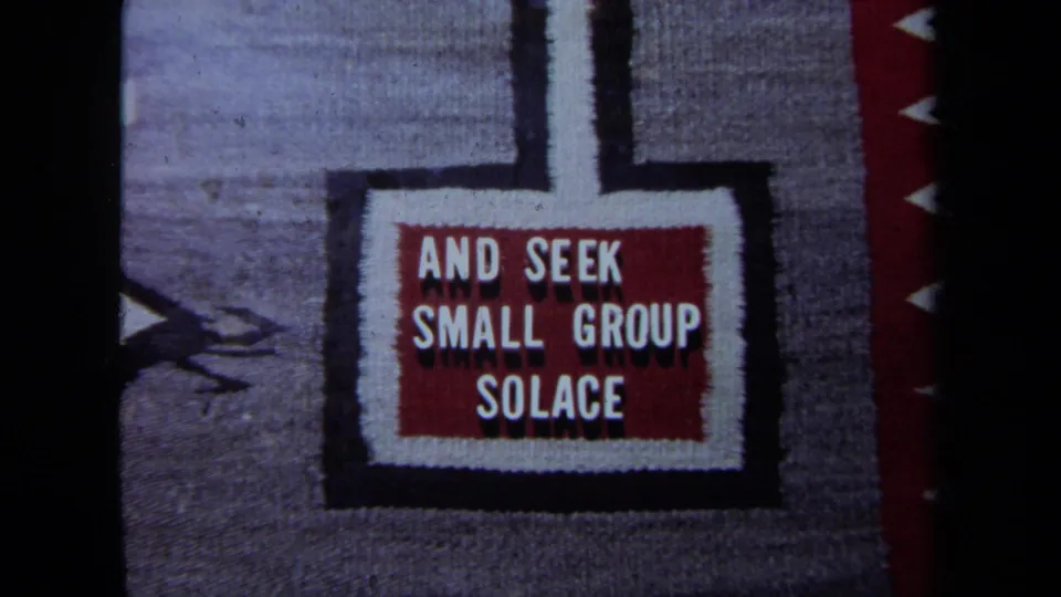 A woven textile sign fills the frame, showing the readable phrase AND SEEK SMALL GROUP SOLACE.