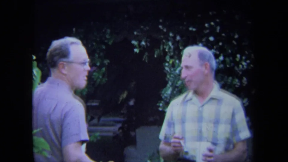 Two adult men in casual 1971 California clothing speak outdoors, with one holding a drink in a leafy garden.