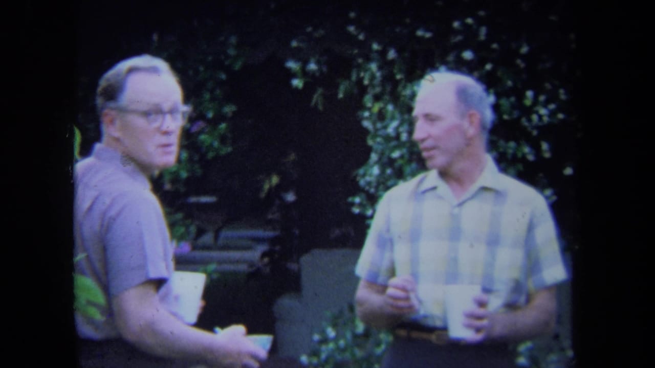 Two adult men stand outdoors holding white cups in a leafy backyard.