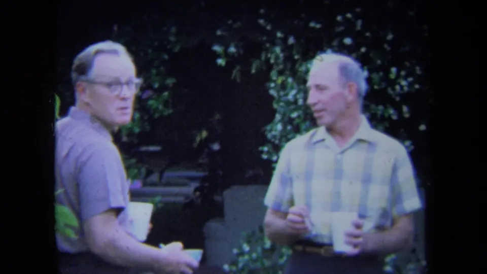 Two adult men in 1971 California home-movie footage stand in a shaded backyard, one looking toward the camera while both hold white cups.