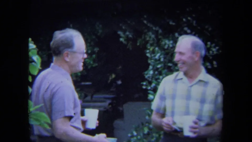 Two adult men in a California backyard appear to chat and smile while holding white cups, framed by dark foliage and patio seating.