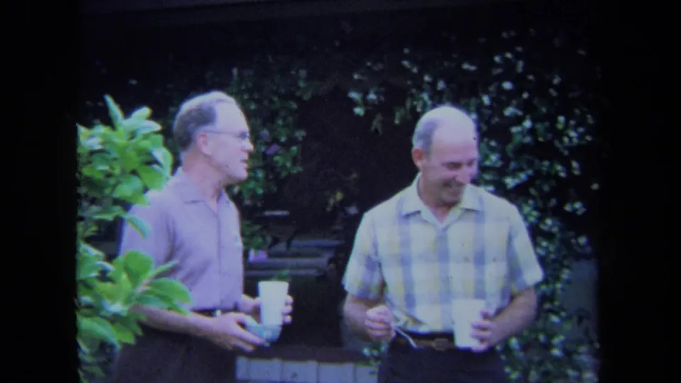 Two adult men in casual 1970s shirts stand near backyard greenery, with one looking down and both holding cups during conversation.