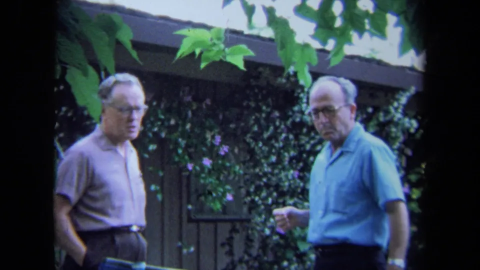 Two adult men in 1971 California backyard footage stand near a wooden fence and leafy branches, one holding a small item or cup.