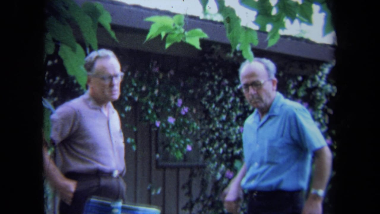 Two older men stand in a leafy backyard garden near a wooden fence.