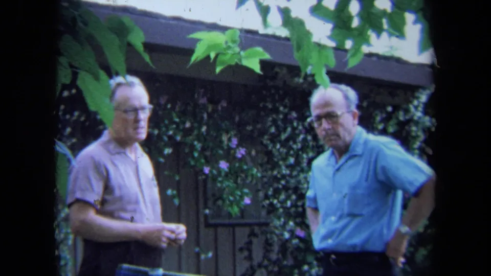 In a 1971 California garden, two older men stand near a wooden fence covered with ivy and small purple flowers.
