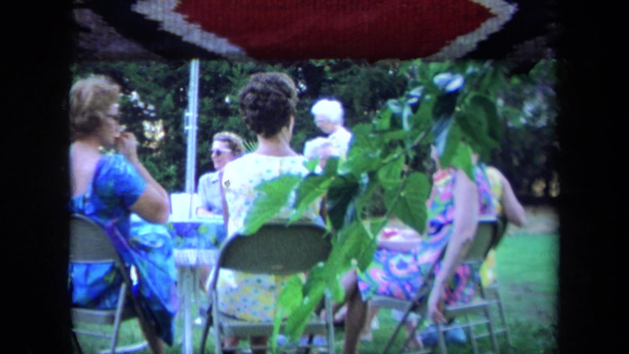 1971 California home-movie frame of women in summer dresses seated on folding chairs under a patio umbrella in a garden.