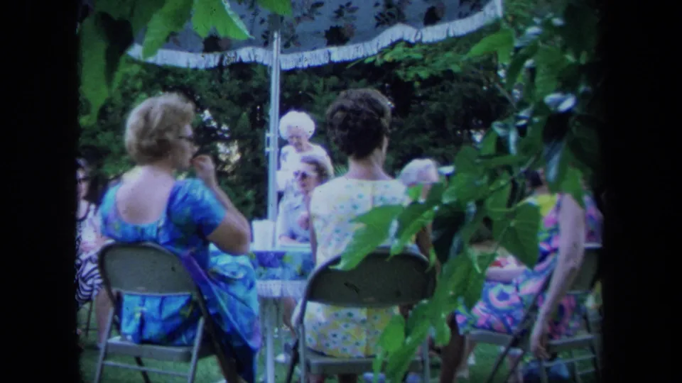 Women at a 1971 California outdoor social gathering sit beneath a fringed umbrella, partly obscured by leafy branches.