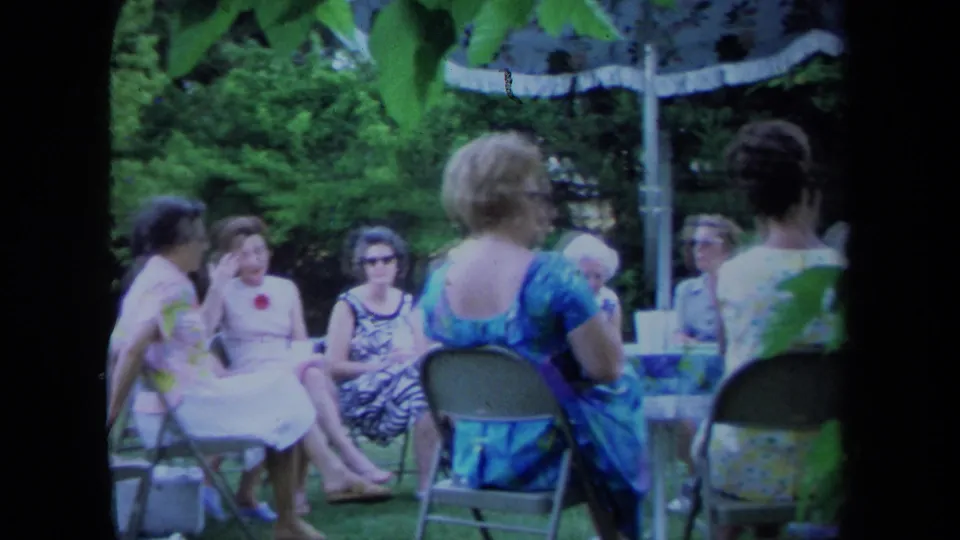 1971 California home-movie frame of women in summer dresses and sunglasses gathered on folding chairs under garden umbrellas.
