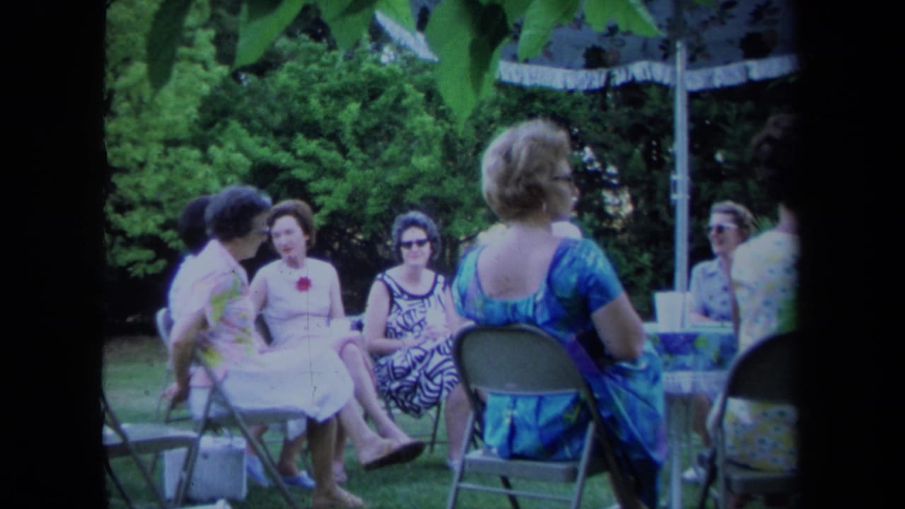 Women sit on folding chairs under a patio umbrella in a leafy yard.