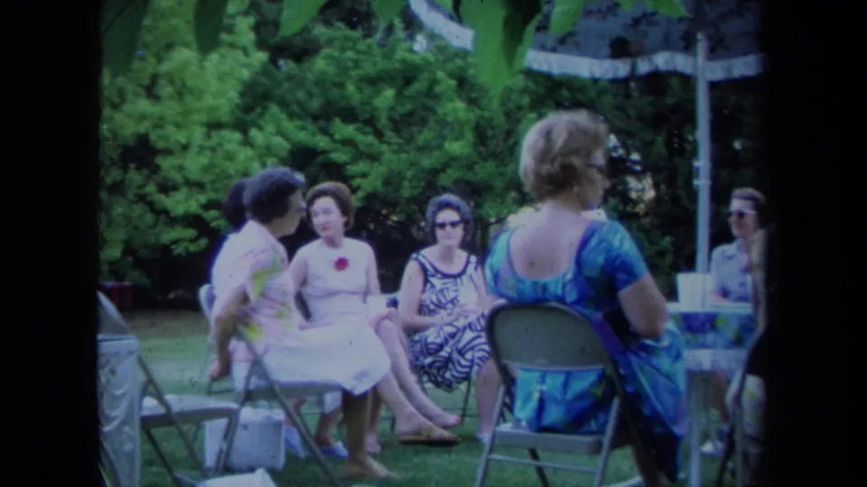 Women in 1970s attire sit together beneath a patio umbrella in a California outdoor home-movie scene.