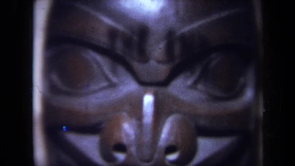 Close-up home-movie frame of a carved wooden mask, softly focused with visible 8mm color grain.
