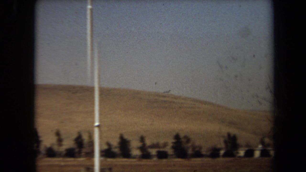 1971 Laguna Hills 8mm frame showing dry hills, hazy sky, and scattered roadside trees viewed from a moving position.
