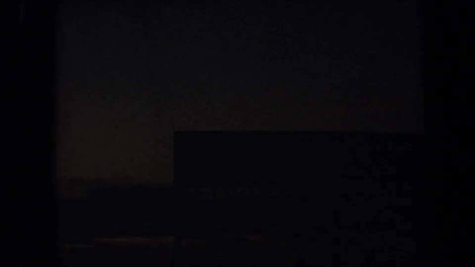 Underexposed 1971 Laguna Hills 8mm frame where a dark rectangular sign or structure sits against the roadside landscape.