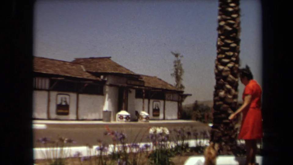 1971 home-movie frame from Laguna Hills showing a woman in a red dress walking a small dog beside flowers and a palm tree near the kennel club building.