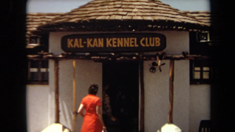 The woman reaches the entrance under the Kal-Kan Kennel Club sign, with the doorway and decorative dog houses visible.