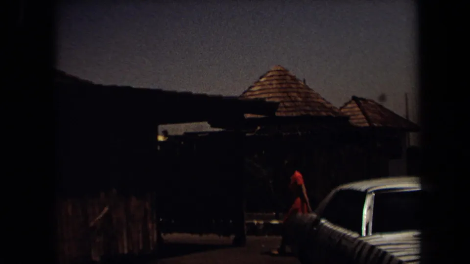 Dim 1971 Laguna Hills 8mm frame with a person in red near rustic fencing, thatched roofs, and a partially visible car.