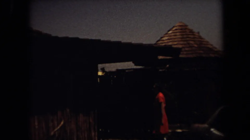 Underexposed 8mm frame of a person in red walking beside dark thatched structures at the Laguna Hills safari site.