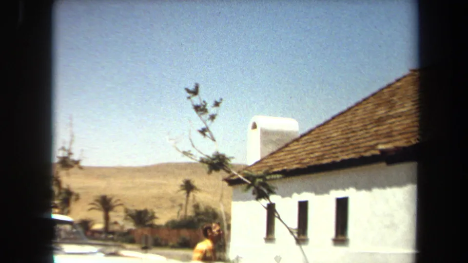 Bright Laguna Hills exterior showing a white stucco building with tiled roof, dry hills, palm trees, and a man partly visible below.