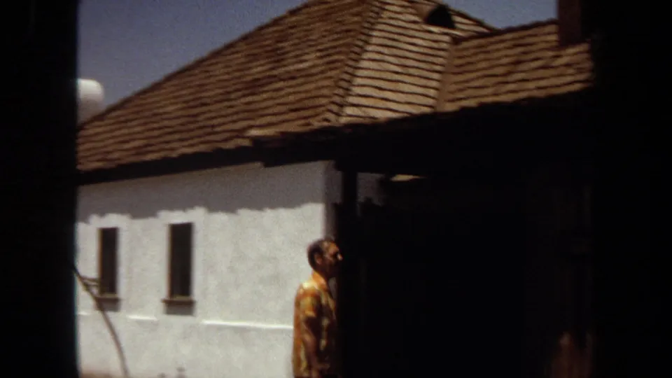 1971 Laguna Hills daytime exterior with a tiled-roof stucco building and a man in a patterned yellow shirt near shade.