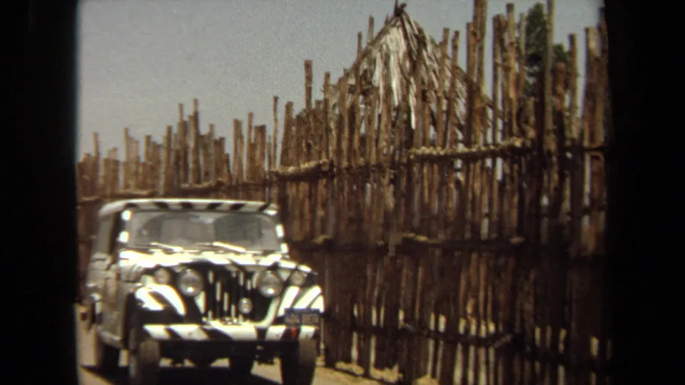 The safari jeep passes nearer to the camera, with its striped hood, license plate area, rough pole fence, and thatched roof detail visible.