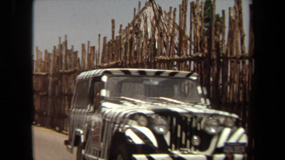 Close Laguna Hills safari-frame view of the zebra-painted jeep as it passes beside vertical pole fencing and a thatched-roof feature.
