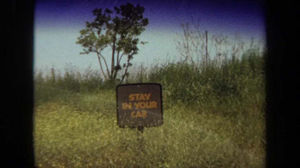 1971 Laguna Hills frame from inside a car showing a weathered sign that reads STAY IN YOUR CAR beside tall grass and trees.