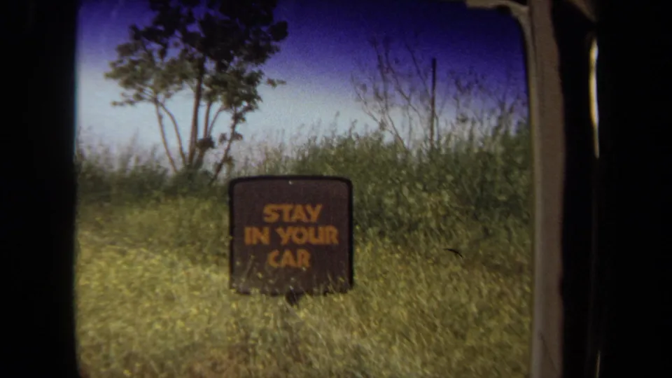 1971 Laguna Hills home-movie frame showing the STAY IN YOUR CAR sign through a car window, with tall grass and trees behind it.
