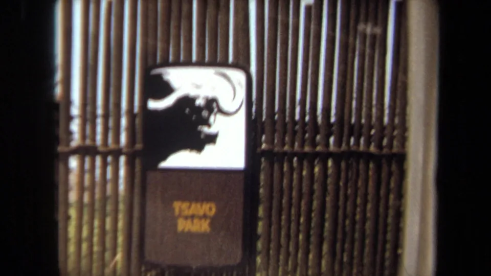 A closer 1971 8mm view shows the Tsavo Park sign and buffalo graphic framed by rustic fencing and a car-window edge.