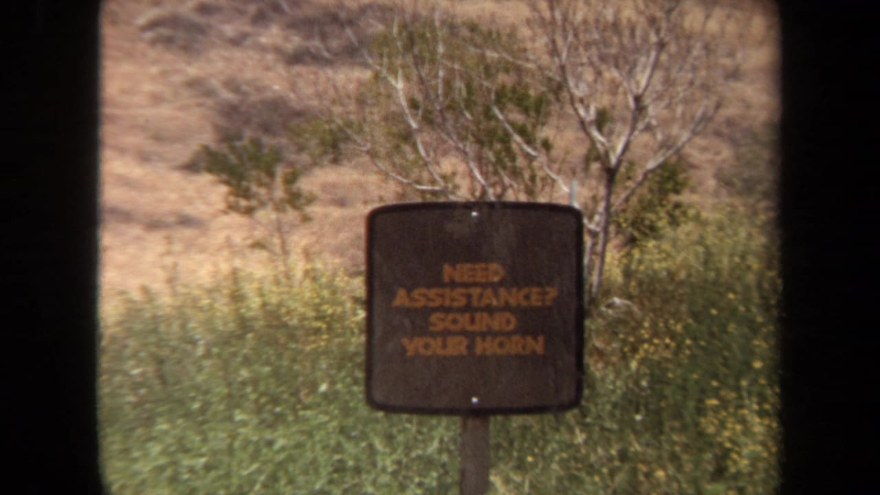 Brown assistance sign standing among dry grass and shrubs in an open landscape.