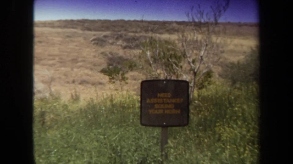 1971 Laguna Hills 8mm frame with the horn-for-assistance sign viewed from farther back through a vehicle window.