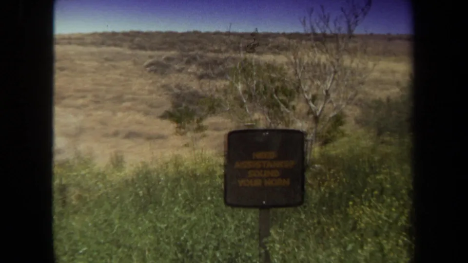 1971 Laguna Hills 8mm frame showing the assistance sign receding in a brushy landscape, framed by the car window and film edge.