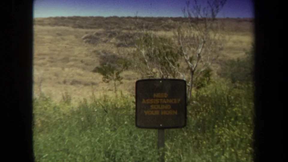 1971 Laguna Hills safari park view from a vehicle, with a small assistance sign, brush, open ground, and dark window edges.