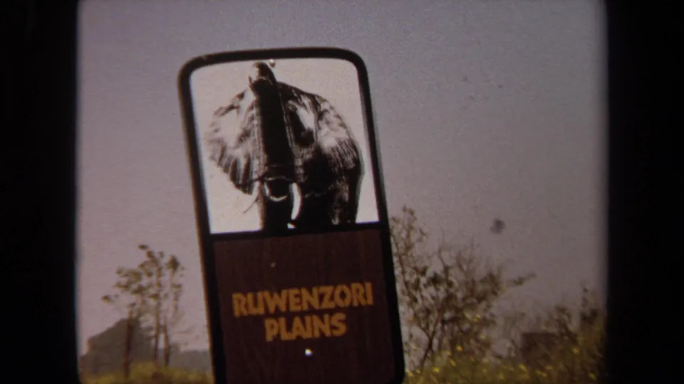 Close 1971 8mm view of the Ruwenzori Plains sign, with an elephant graphic above yellow lettering and sparse roadside trees.