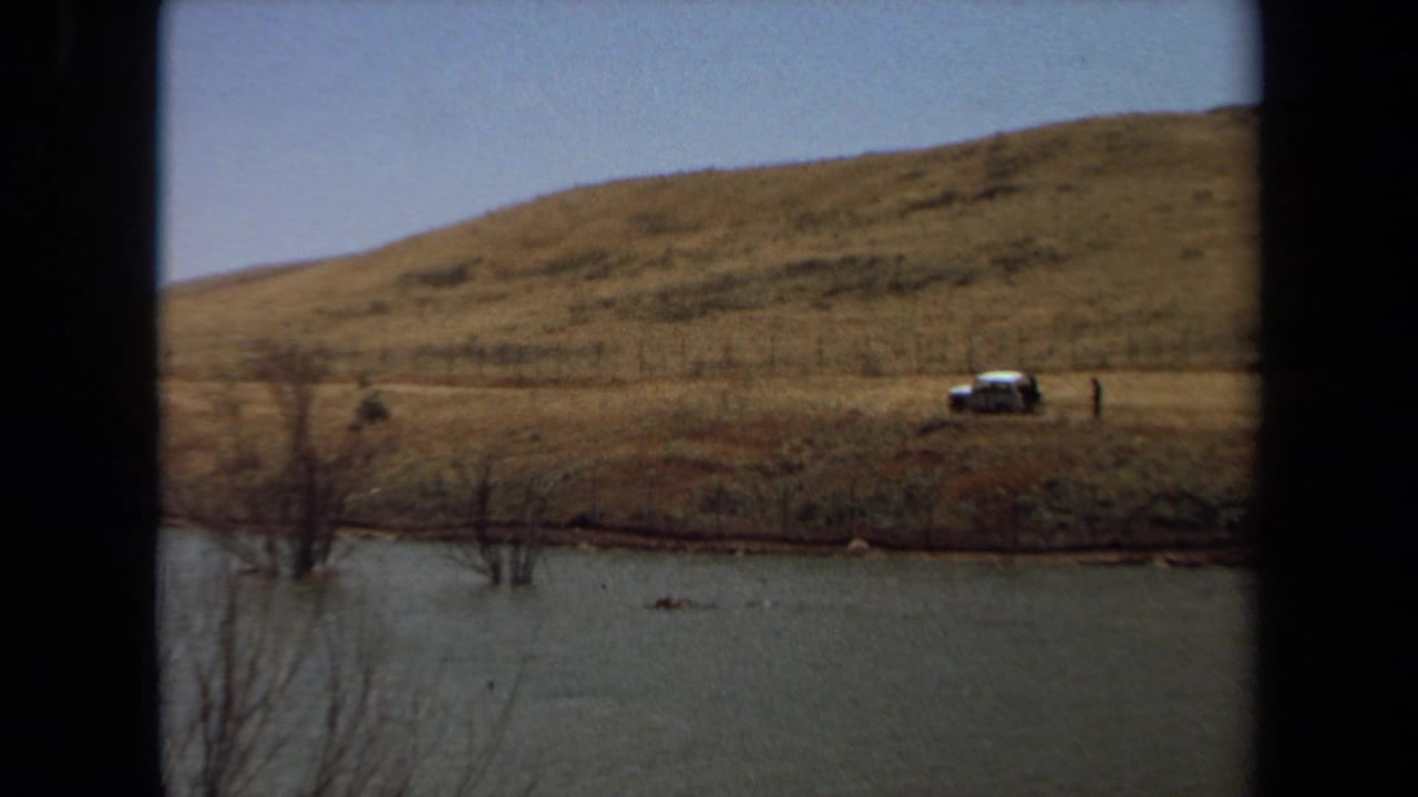 1971 Laguna Hills 8mm frame showing a parked car on a grassy hillside overlooking a pond and fenced shore.